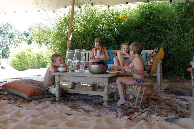 Gezelschap aan een lage tafel met kussens in het zand onder een luifel in de tuin
