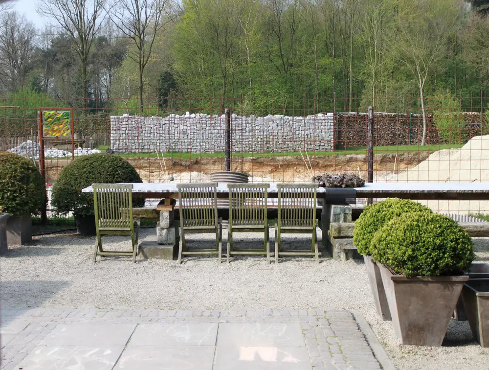 Lange eettafel met stoelen en geschoren vormen op een terras, met bouwwerk erachter